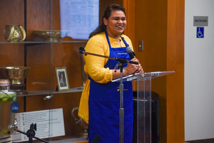 Woman in yellow shirt and apron speaks on stage.