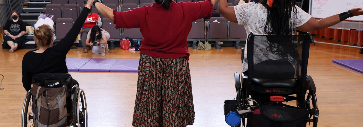 Three disabled dancers touching hands with arms raised; wheelchair user (left), person standing (center) and person standing with a walker (right)