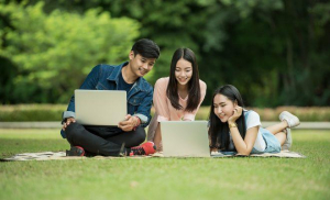A group of students sit and lay on a field looking at a laptop.