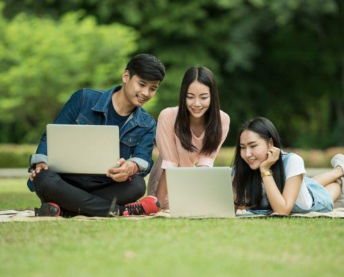 A group of students sit and lay on a field looking at a laptop.