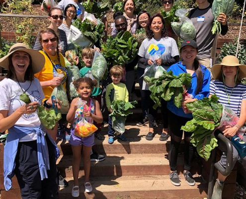 A group of volunteers of all ages including children holds fresh vegetables.