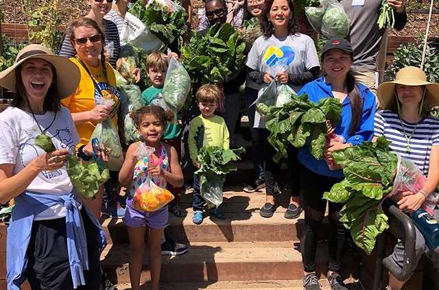 A group of volunteers of all ages including children holds fresh vegetables.
