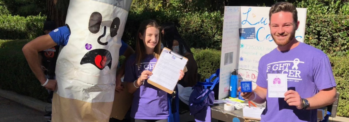 2 people standing in front of a board promotes the fight for lunch cancer with a cigarette mascot.