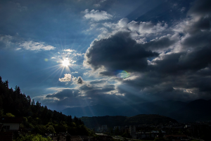 A blue sky has a sun peaking through the clouds overlooking the forest.