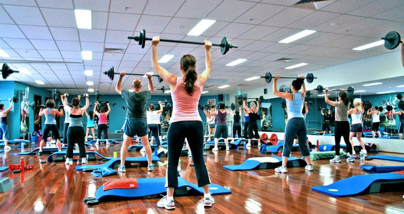 A room full of people practice weightlifting by lifting barbells above their heads.