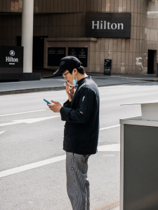 Man stands on the side of the road in front of the Hilton smoking a cigarette with a mask on.