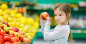 A young girl taps on an apple in the produce aisle of a grocery store.