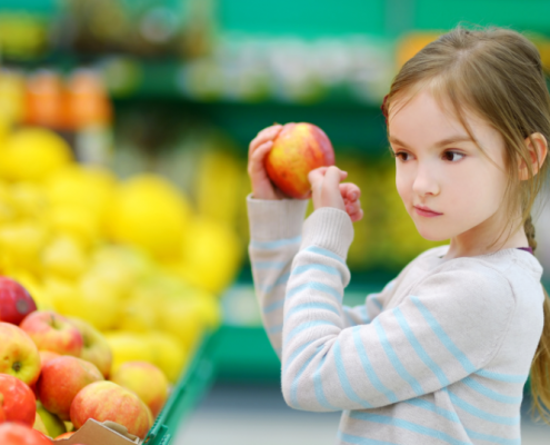 A young girl taps on an apple in the produce aisle of a grocery store.