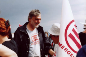 A man stands in a crowd wearing an emerge shirt next to an emerge flag.