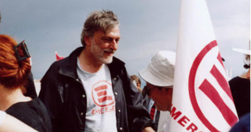A man stands in a crowd wearing an emerge shirt next to an emerge flag.
