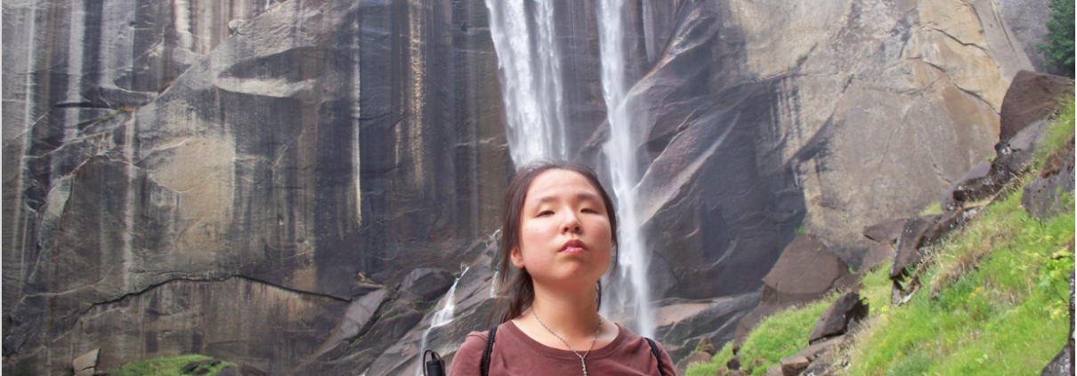 A girl poses in front of a waterfall holding a white cane.