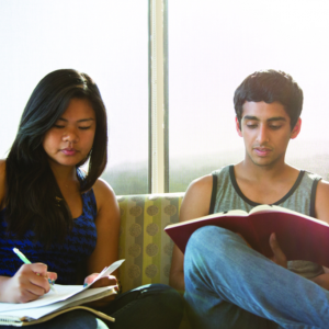 2 students sit next to one another, one that Is writing notes and the other reading.