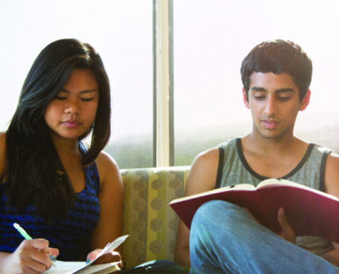 2 students sit next to one another, one that Is writing notes and the other reading.
