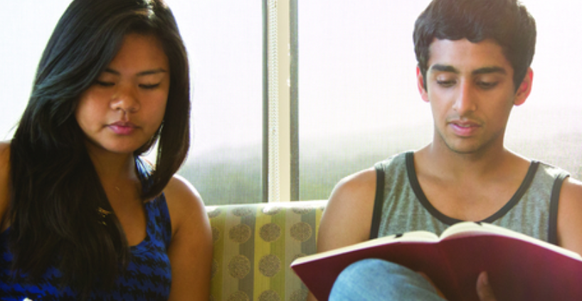 2 students sit next to one another, one that Is writing notes and the other reading.