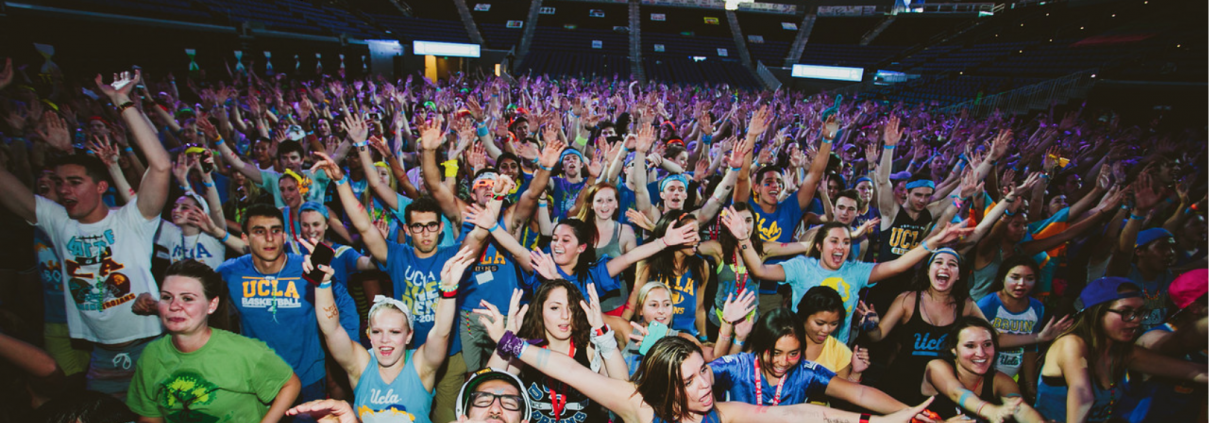 A very large crowd of students hold both of their arms up and out.