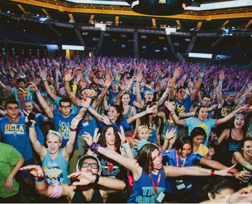 A very large crowd of students hold both of their arms up and out.