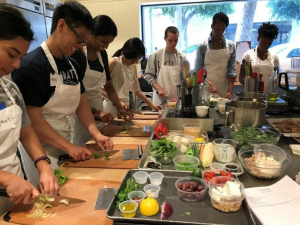 A group of people stand around a kitchen cutting up vegetables and cooking with one another.