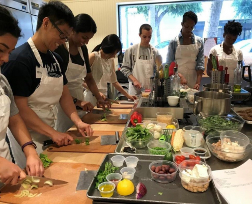 A group of people stand around a kitchen cutting up vegetables and cooking with one another.