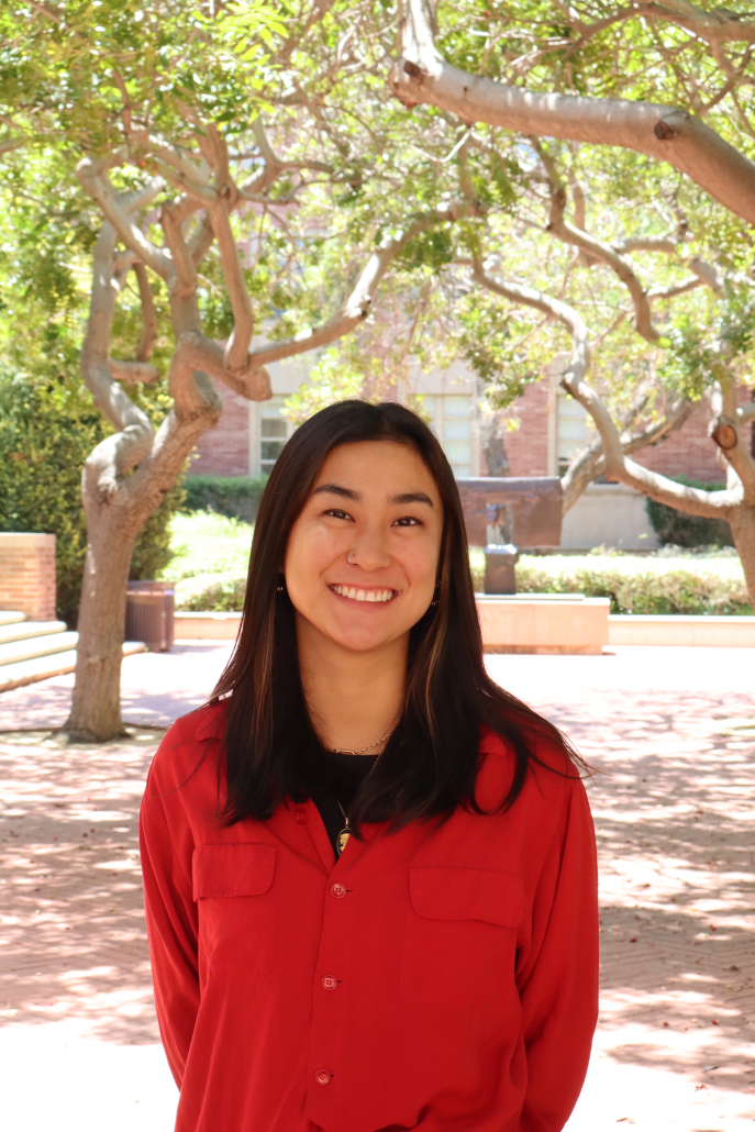 Photo depicts Ikuko "Coco" Nakano in a red button down shirt. Background depicts Murphy Hall courtyard at UCLA with trees and dappled sunlight.