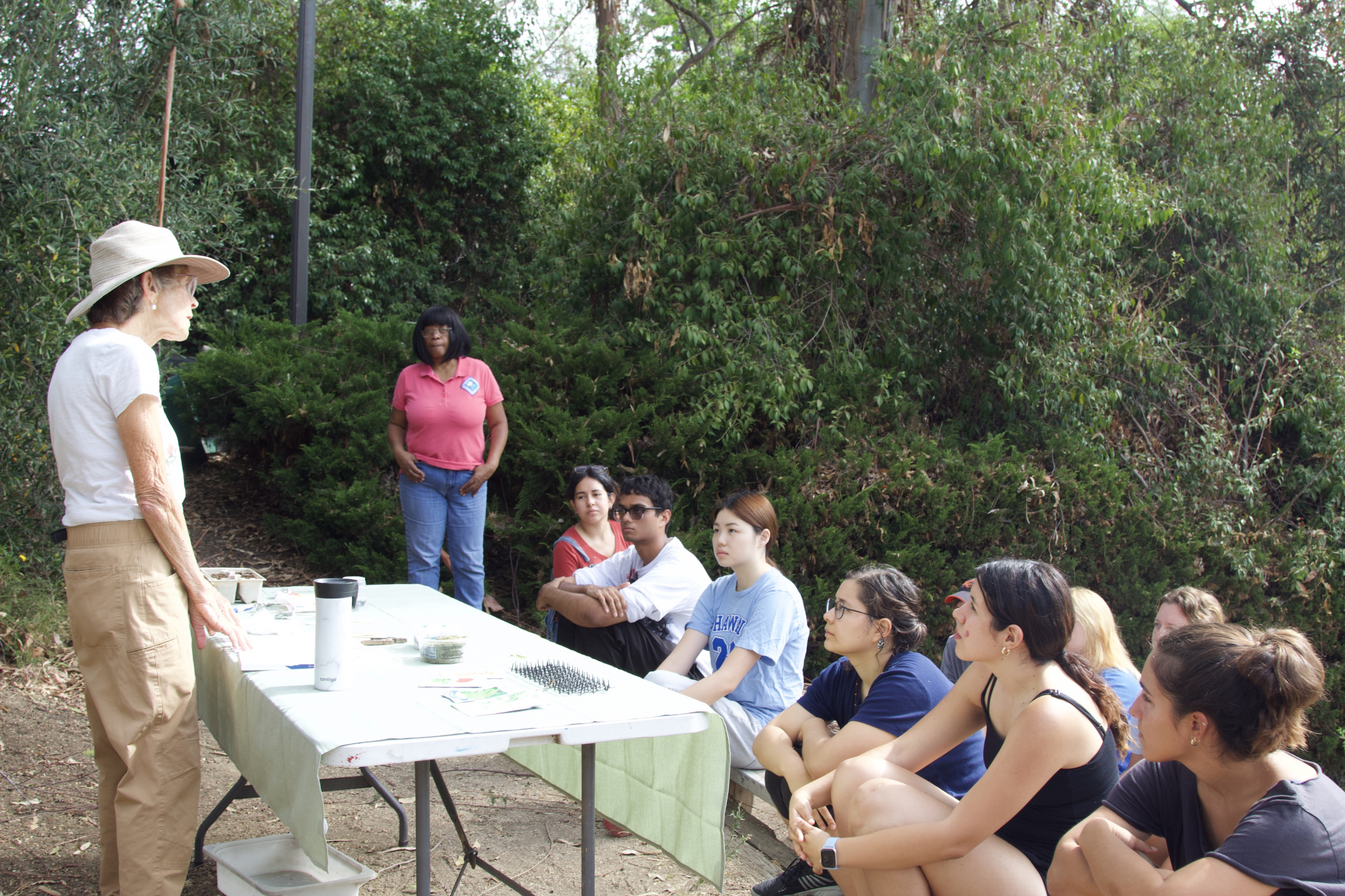 Master Gardener teaches introductory gardening principles to 9 pictured workshop attendees. They are separated by a table which has seed packets and other gardening supplies for demonstration.