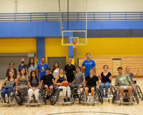 16 individuals posed on a basketball court in two rows. The back row are standing and the front row are sitting in wheelchairs. Two individuals are holding a basketball.