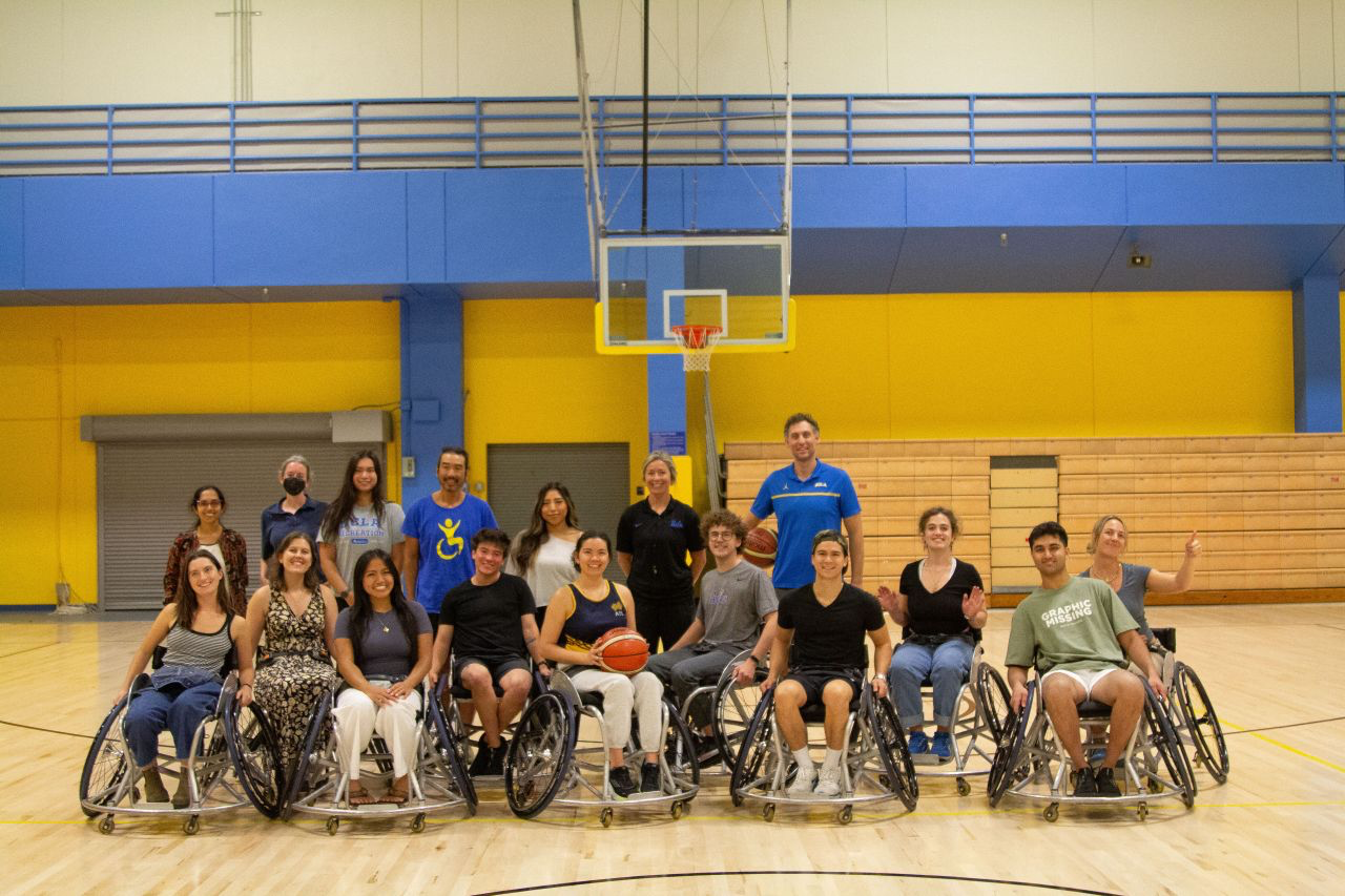 16 individuals posed on a basketball court in two rows. The back row are standing and the front row are sitting in wheelchairs. Two individuals are holding a basketball.